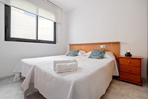 A neatly made bed with a white bedspread and two patterned pillows, a wooden nightstand with a lamp, and a window with blinds.
