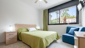 A bedroom with a bed, a dresser, a blue sofa, and a window with a view of palm trees and buildings.