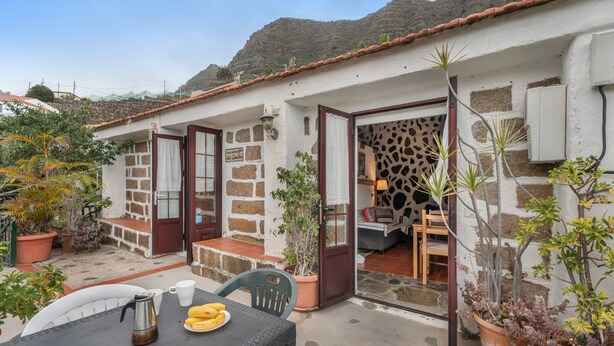 Une petite maison avec un patio, une table sur laquelle se trouvent une cafetière et des bananes, ainsi que des plantes en pot.