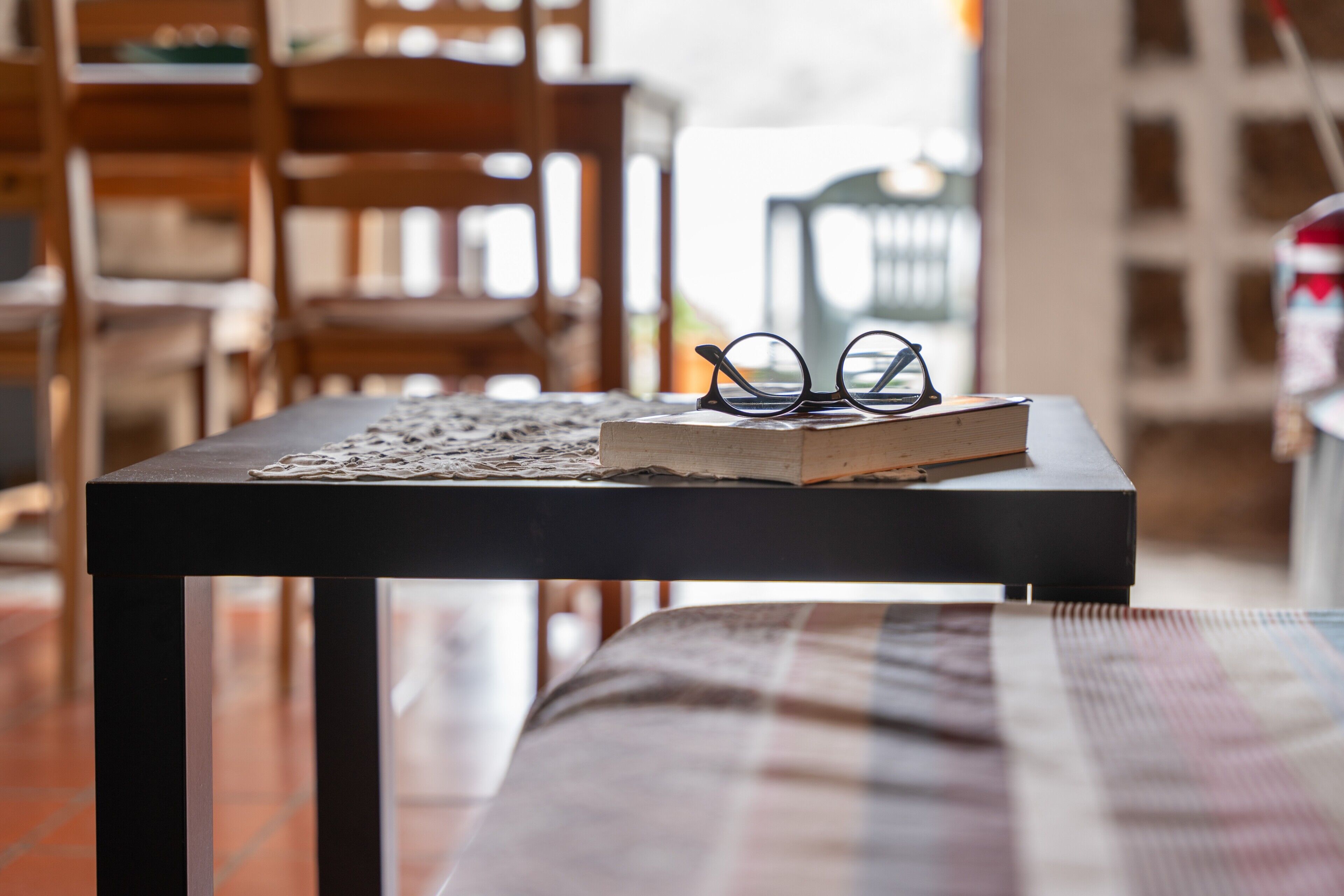 A table with a book and glasses on it, a plaid blanket on a bench, and wooden chairs in the background.