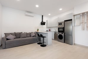 A modern kitchen with a grey sofa, two black bar stools, a built-in island, a washing machine, and a stainless steel refrigerator.
