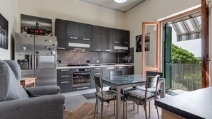 A modern kitchen with dark cabinetry, a dining area with a glass table, and a view of greenery through an open door. - House of Flowers holiday home with balcony and Wi-Fi (Montopoli vald’arno)