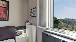A bedroom with a bed, bedside table, radiator, and a window with a view of a forest. - House of Flowers holiday home with balcony and Wi-Fi (Montopoli vald’arno)