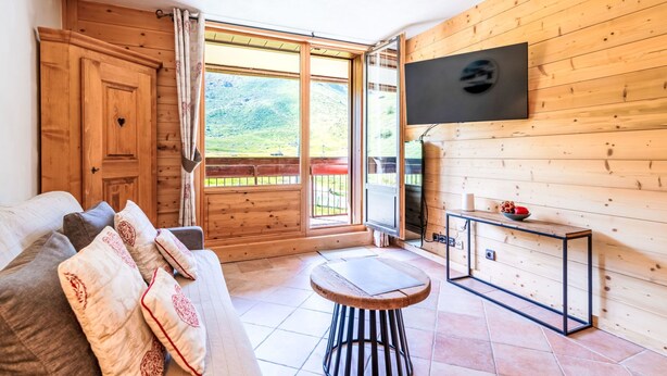 A cozy room with a wooden interior, a sofa with patterned cushions, a small round table, a TV mounted on the wall, and a large window offering a view of a green landscape.