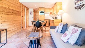 A cozy room with a wooden wall, a dining area with a table and chairs, a sofa with decorative pillows, and a kitchen in the background.