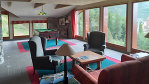 Une pièce équipée d’une table de billard, de chaises, d’un canapé, d’une lampe et de grandes fenêtres donnant sur un paysage verdoyant et des bâtiments.