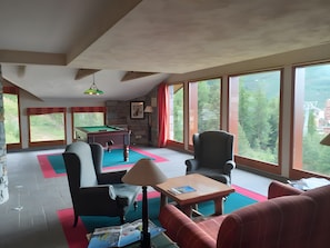 Une pièce équipée d’une table de billard, de chaises, d’un canapé, d’une lampe et de grandes fenêtres donnant sur un paysage verdoyant et des bâtiments.