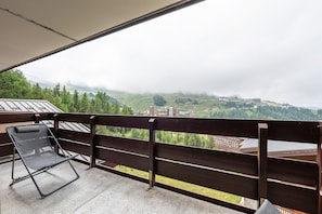 Un balcon doté d’une chaise à ossature métallique et d’une balustrade en bois, offrant une vue sur un paysage vallonné parsemé de bâtiments.