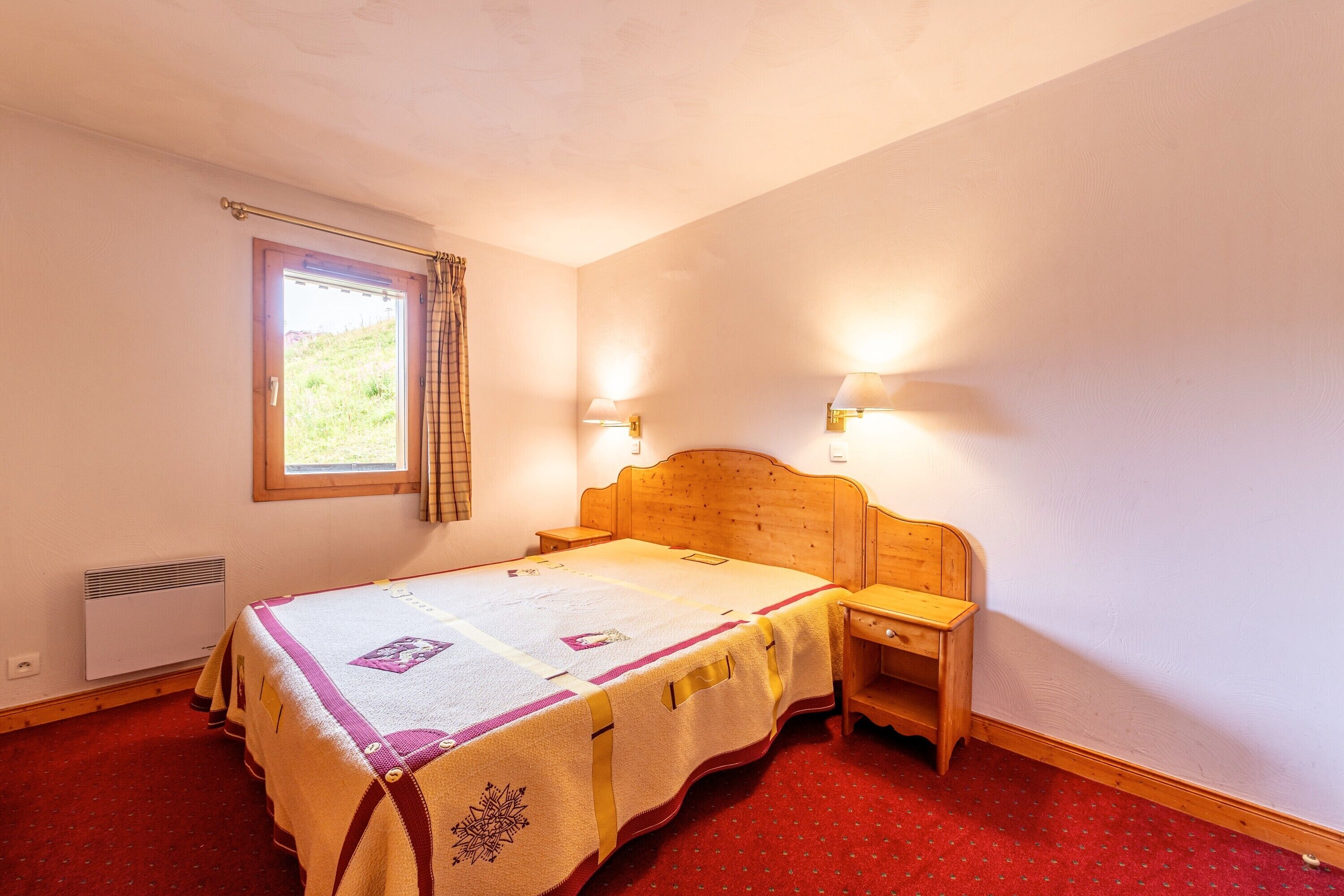 A bedroom with a wooden bed, a window with a curtain, two bedside lamps, and a red carpet.