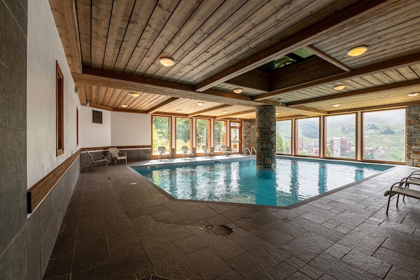 Une piscine intérieure agrémentée de piliers en pierre, de poutres en bois et de grandes fenêtres offrant une vue sur l’extérieur.