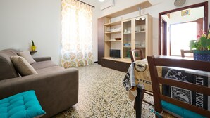 Ein Wohnzimmer mit einer Couch, einem Fernseher, einem Schrank mit Regalen und einem Essbereich mit einem Stuhl und Tisch.