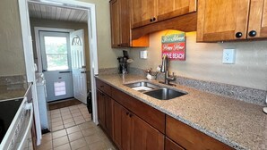 Une cuisine avec des meubles de rangement en bois, un plan de travail en granit, un évier et une porte donnant sur une autre pièce.