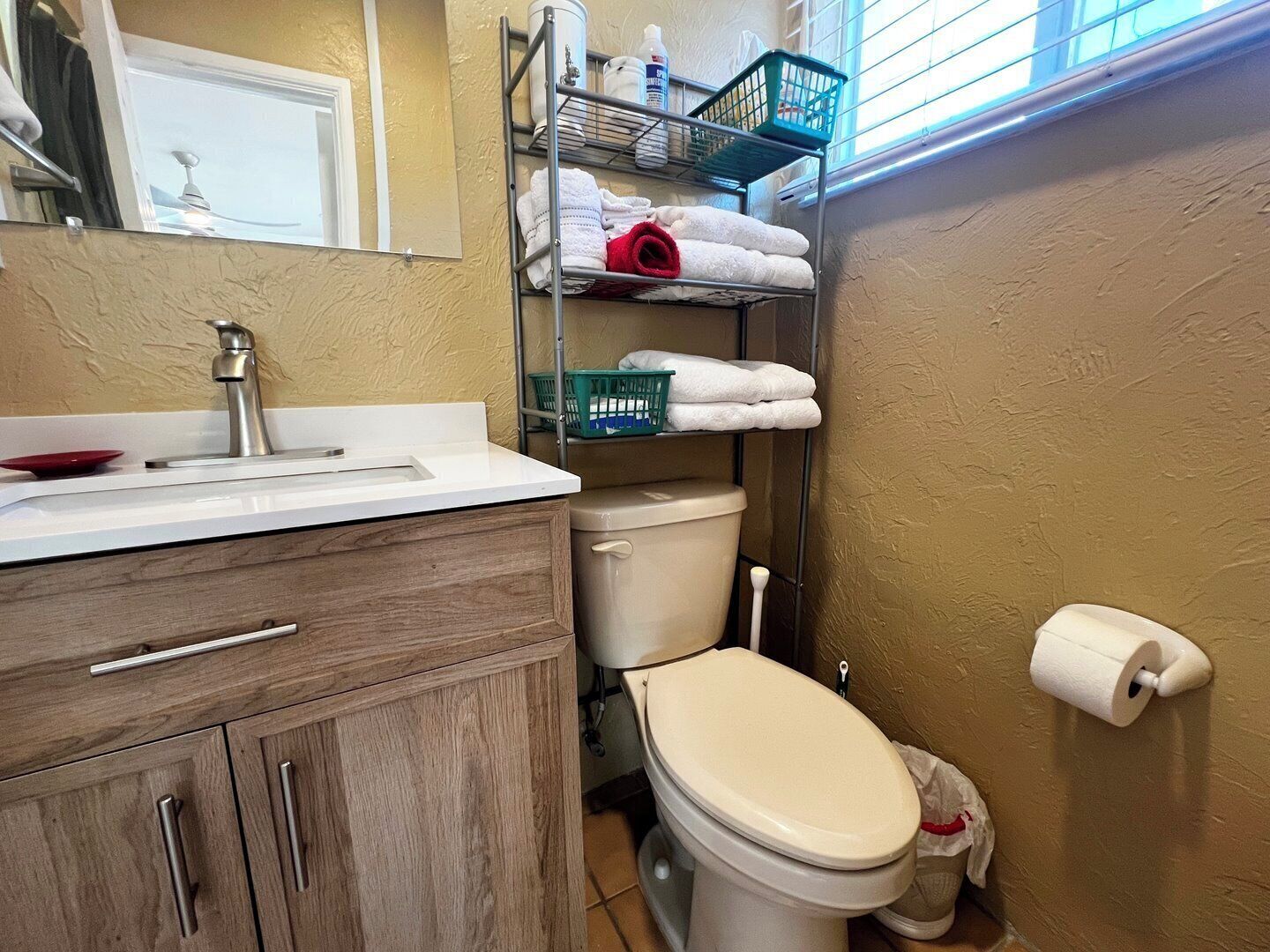 Un baño con un lavamanos blanco, un mueble de madera y un inodoro. Hay repisas con toallas y artículos de aseo.