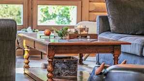Un salon chaleureux avec une table basse en bois, un canapé bleu et une vue sur la verdure grâce à de grandes fenêtres.