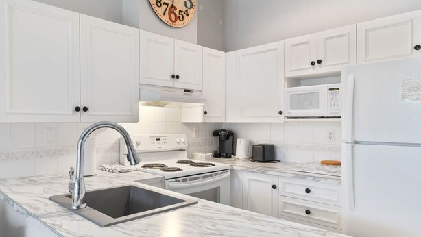 A modern kitchen with white cabinets, a stainless steel sink, a stove, a microwave, and a refrigerator. - Fairway Haven - #104A (Kelowna)