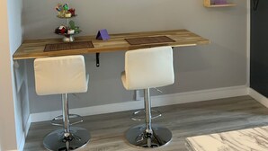 A modern kitchen with a wooden table, two white bar stools, and a decorative wall panel with flowers. - Fairway Haven - #104A (Kelowna)