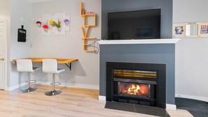 A modern living room with a fireplace, a TV mounted above it, and a wooden table with bar stools. - Fairway Haven - #104A (Kelowna)