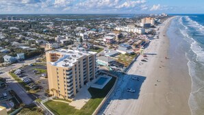 An aerial view of a coastal city with a beachfront hotel featuring multiple floors, a swimming pool, and a parking area. - Oceanfront Condo at Oceania Beach Club - Pool, Balcony, Sleeps 4, Walk to Flagler Ave! (New Smyrna Beach)