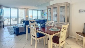 A dining area with a wooden table and chairs, a ceiling fan, and a large mirror. - Oceanfront Condo at Oceania Beach Club - Pool, Balcony, Sleeps 4, Walk to Flagler Ave! (New Smyrna Beach)