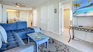 A living room with a sectional sofa, a coffee table, a TV, and a kitchen in the background. - Oceanfront Condo at Oceania Beach Club - Pool, Balcony, Sleeps 4, Walk to Flagler Ave! (New Smyrna Beach)
