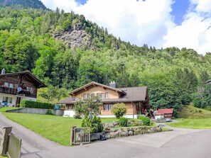 Een chalet met een schuin dak, omringd door weelderige begroeiing en een bergachtig landschap.