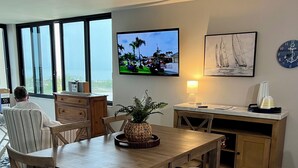 A modern living room with a wooden dining table, striped bench, and a television displaying a tropical scene. - FOUND! Million Dollar View! Stunning Fort Meyers Beach Condo! Book Early & Save! (Fort Myers Beach)