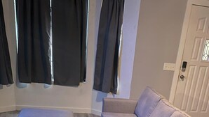 Une pièce avec un canapé beige et un pouf assorti, une fenêtre avec des rideaux noirs et une porte blanche.