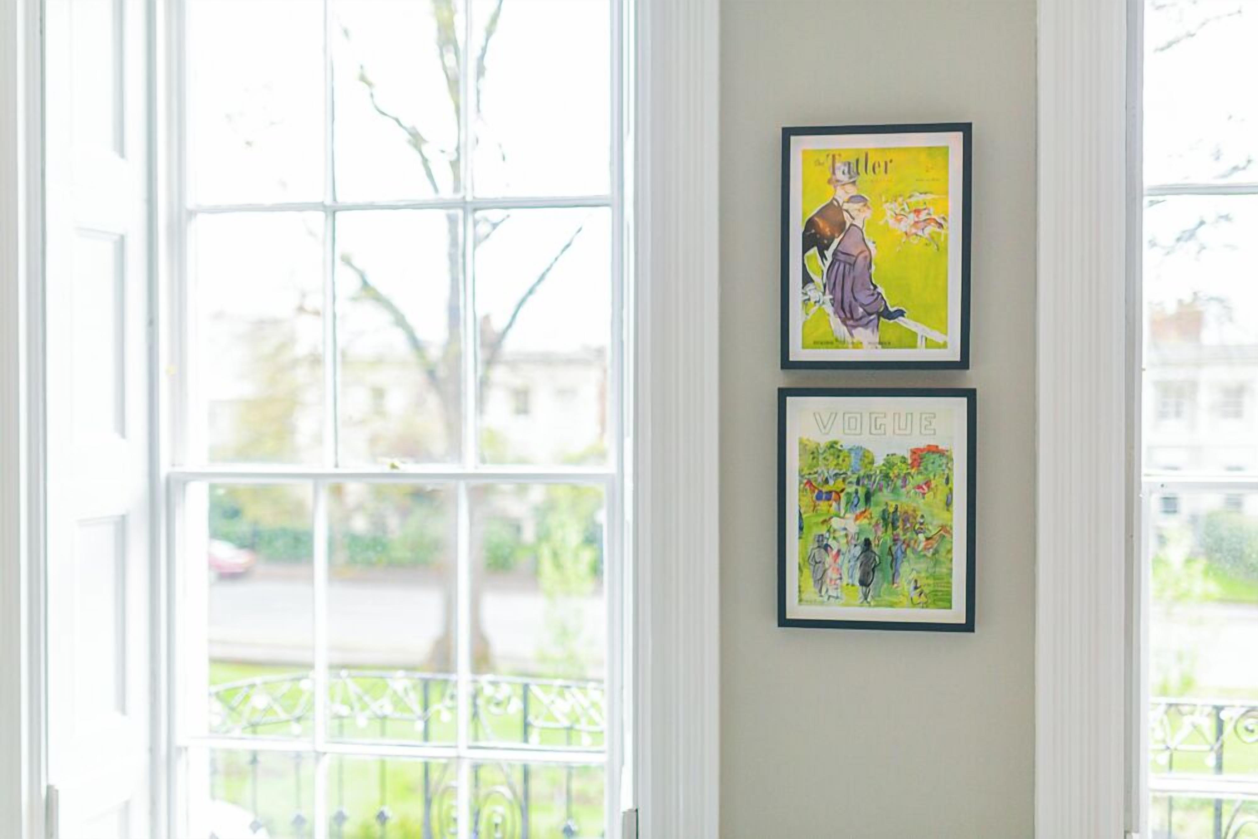 A room with large windows, two framed artworks on the wall, and a view of a street and greenery outside.