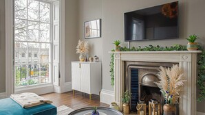 A living room with a fireplace, a sofa, a TV, and a window with a view of buildings. - Stylish Grade II Apartment - The London (Cheltenham)