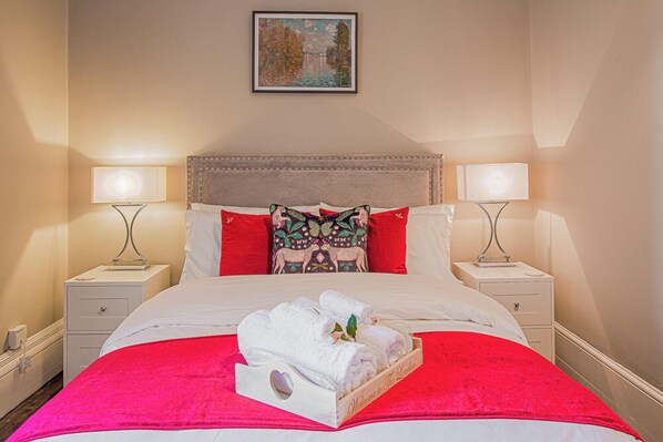 A neatly made bed with white linens, a red throw blanket, and a wooden tray with folded towels. There are two bedside lamps, a framed painting on the wall, and a small nightstand with a lamp.