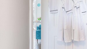 A hotel room with white robes hanging on a door, a blue chair, and a small table with flowers. - Stylish Grade II Apartment - The London (Cheltenham)