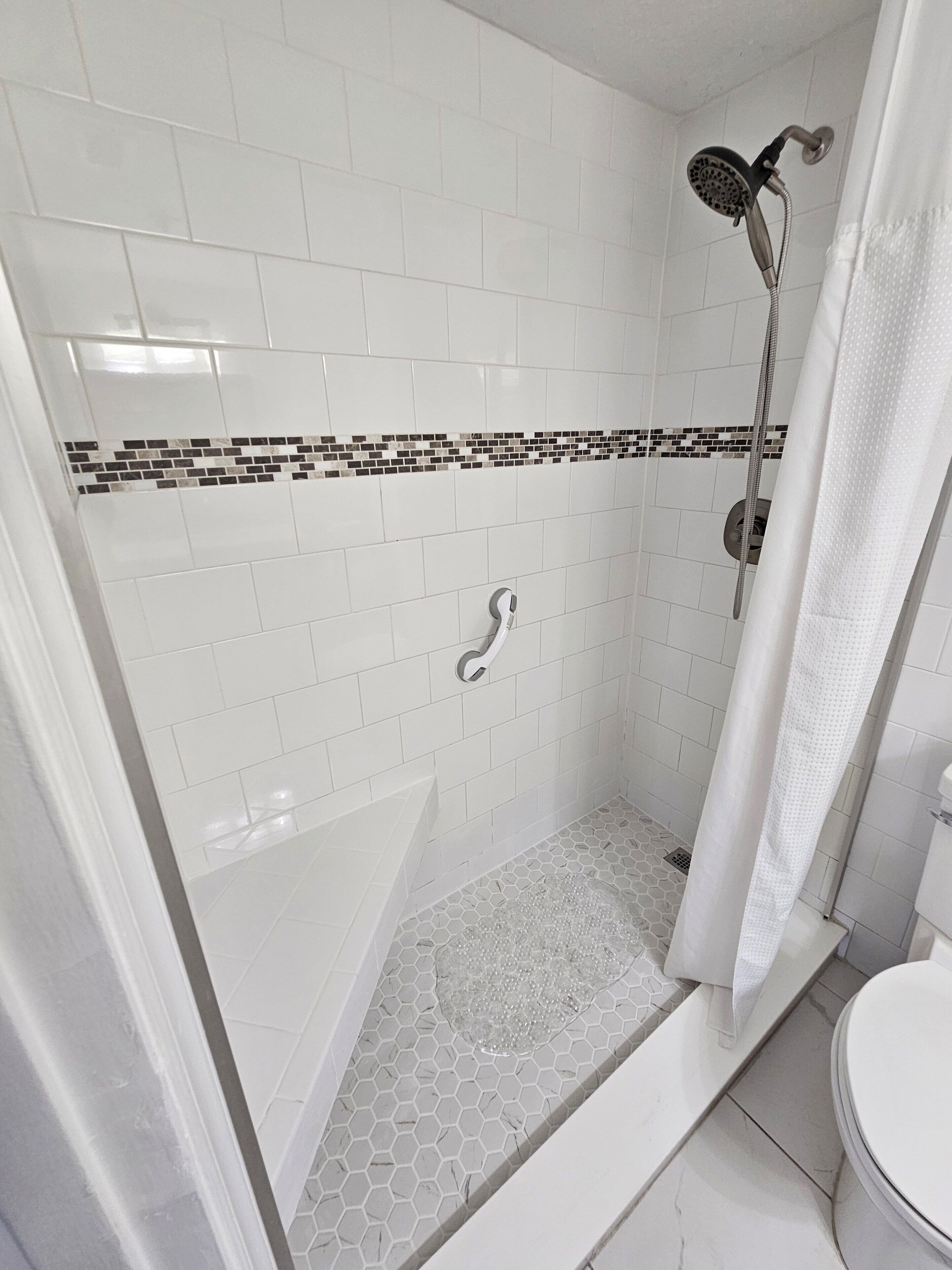 Un bagno con pareti piastrellate in bianco, una doccia con soffione manuale e un pavimento a mosaico esagonale.