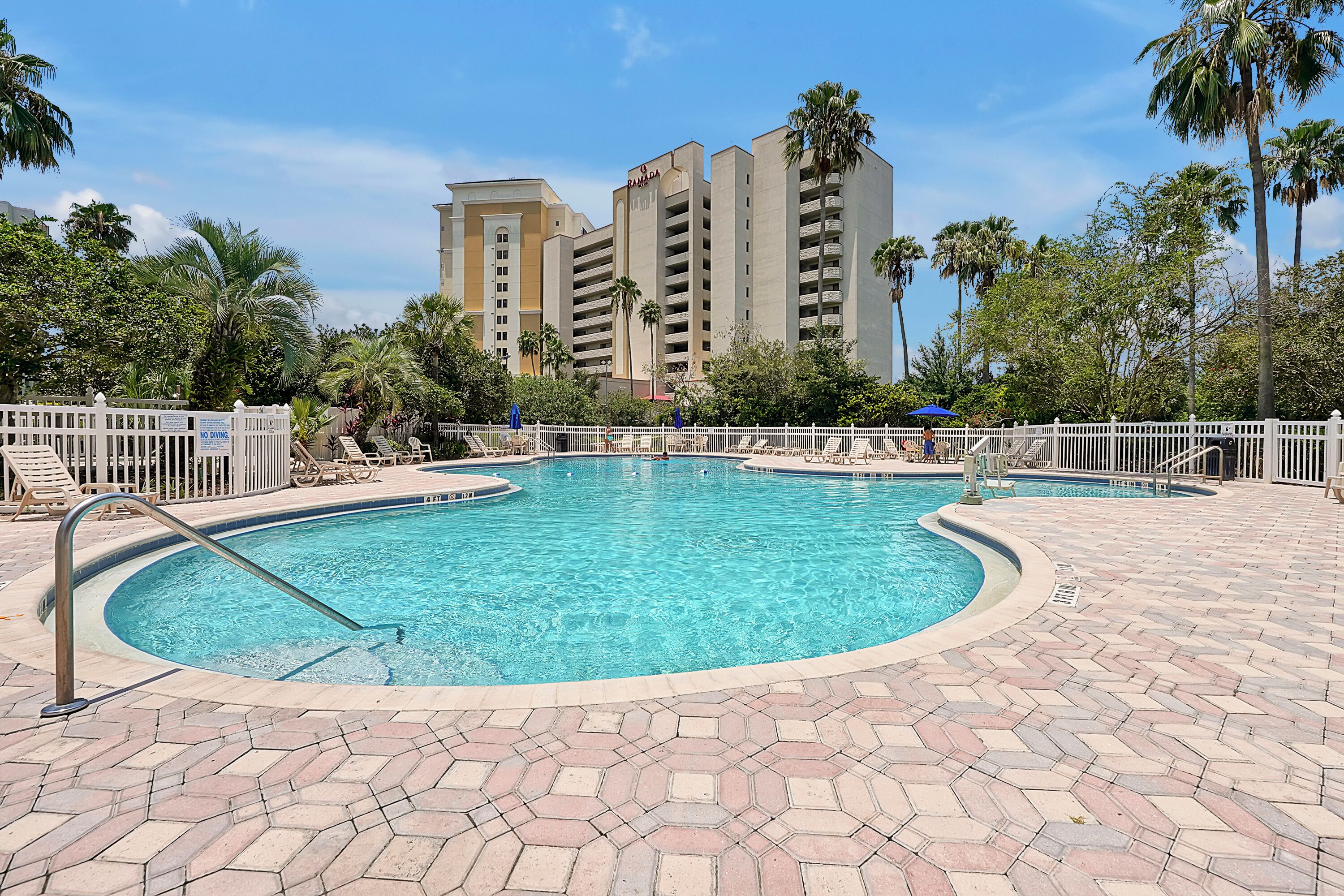A swimming pool with a white fence and lounge chairs, surrounded by palm trees and a hotel building in the background.