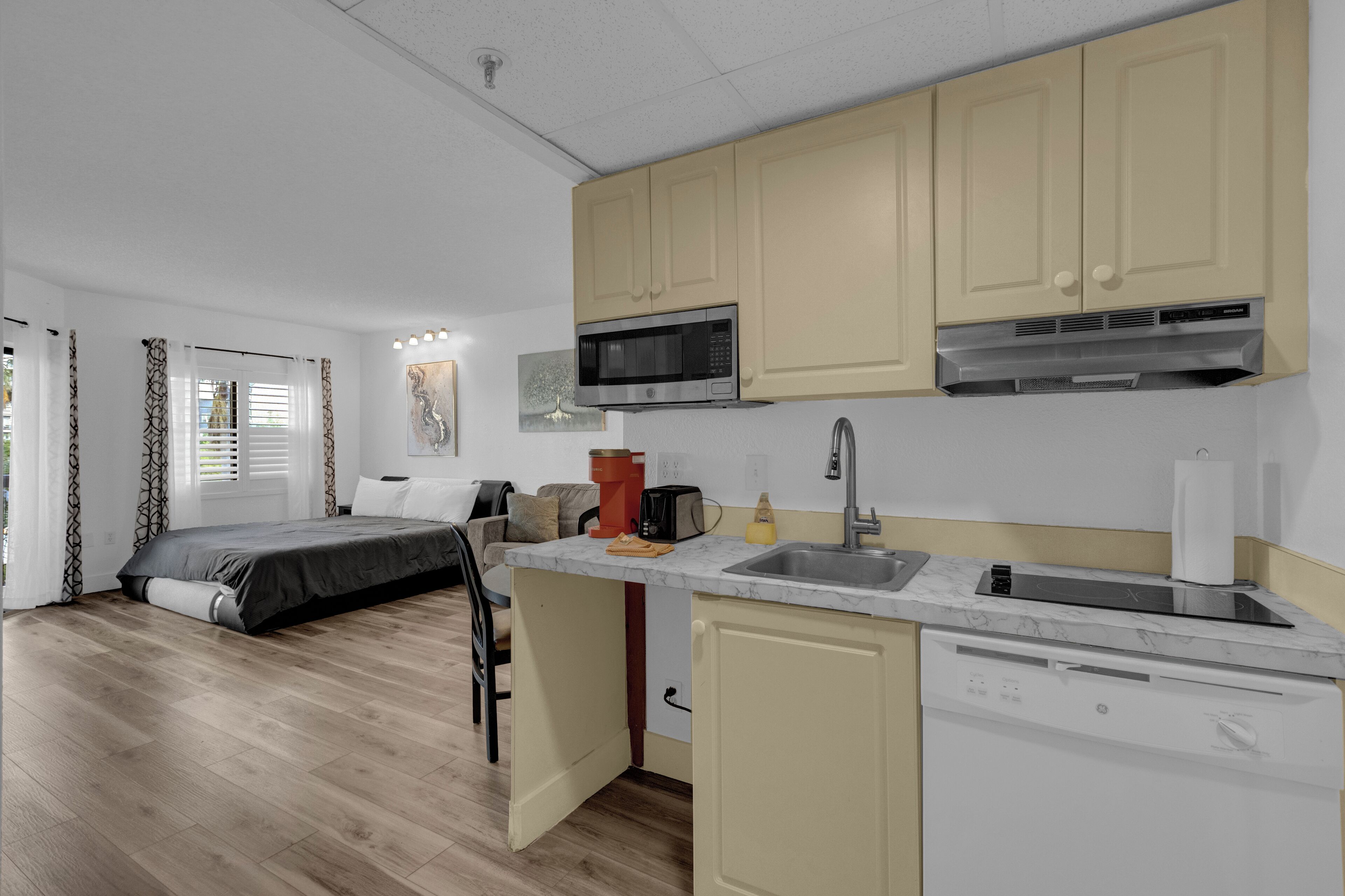 A compact kitchen with a microwave, sink, and dishwasher. A bed and a chair are visible in the adjacent room.
