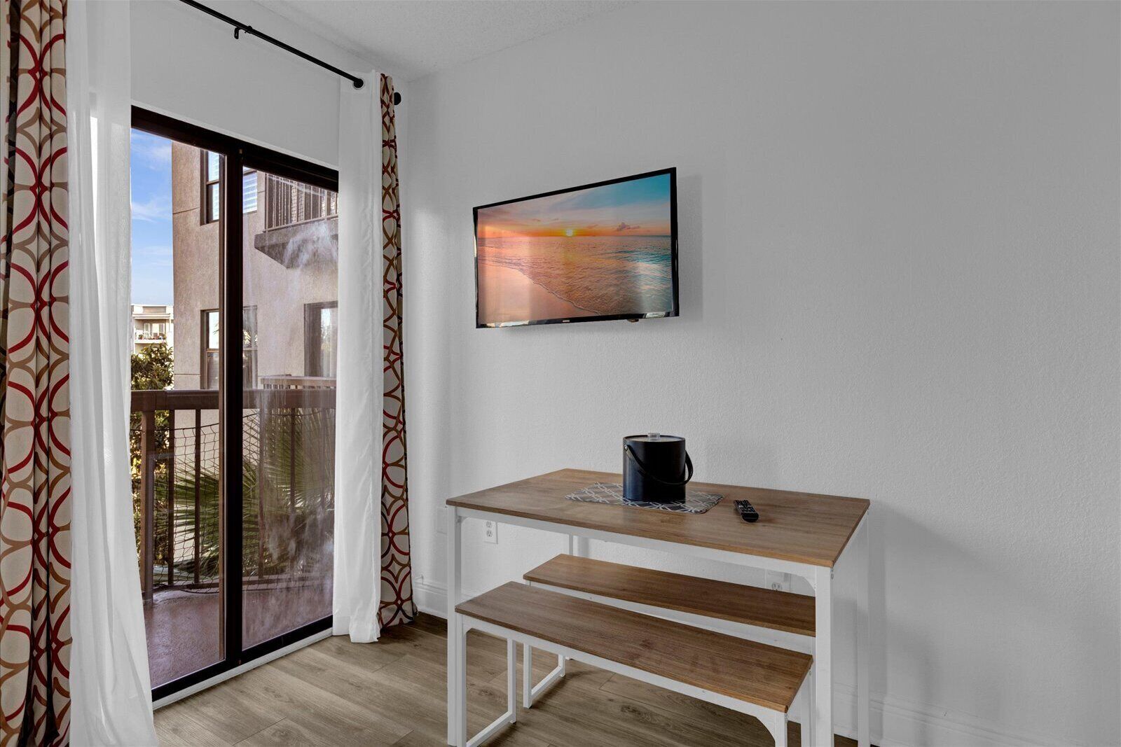 A room with a wooden table, a TV mounted on the wall, and a sliding glass door leading to an outdoor balcony.