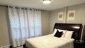 A bedroom with a bed, a nightstand, a floor lamp, a bench, and a window with curtains. - Newly Renovated Luxury 2-BD 2-BA house, AC, WiFi. Mins to NYC, EWR, Newark Distr (Union)