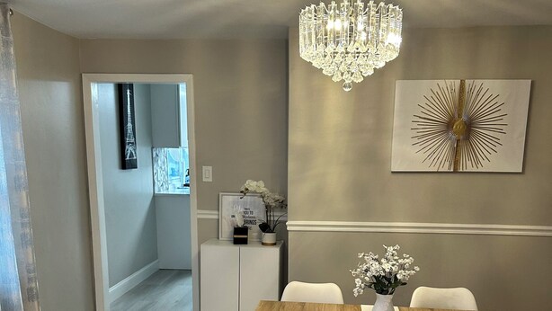 A dining area with a wooden table and white chairs, a chandelier, and a wall art piece. - Newly Renovated Luxury 2-BD 2-BA house, AC, WiFi. Mins to NYC, EWR, Newark Distr (Union)