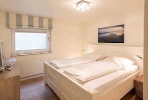 Un dormitorio con cama, ventana con persianas, mesita de noche, lámpara y un cuadro en la pared.