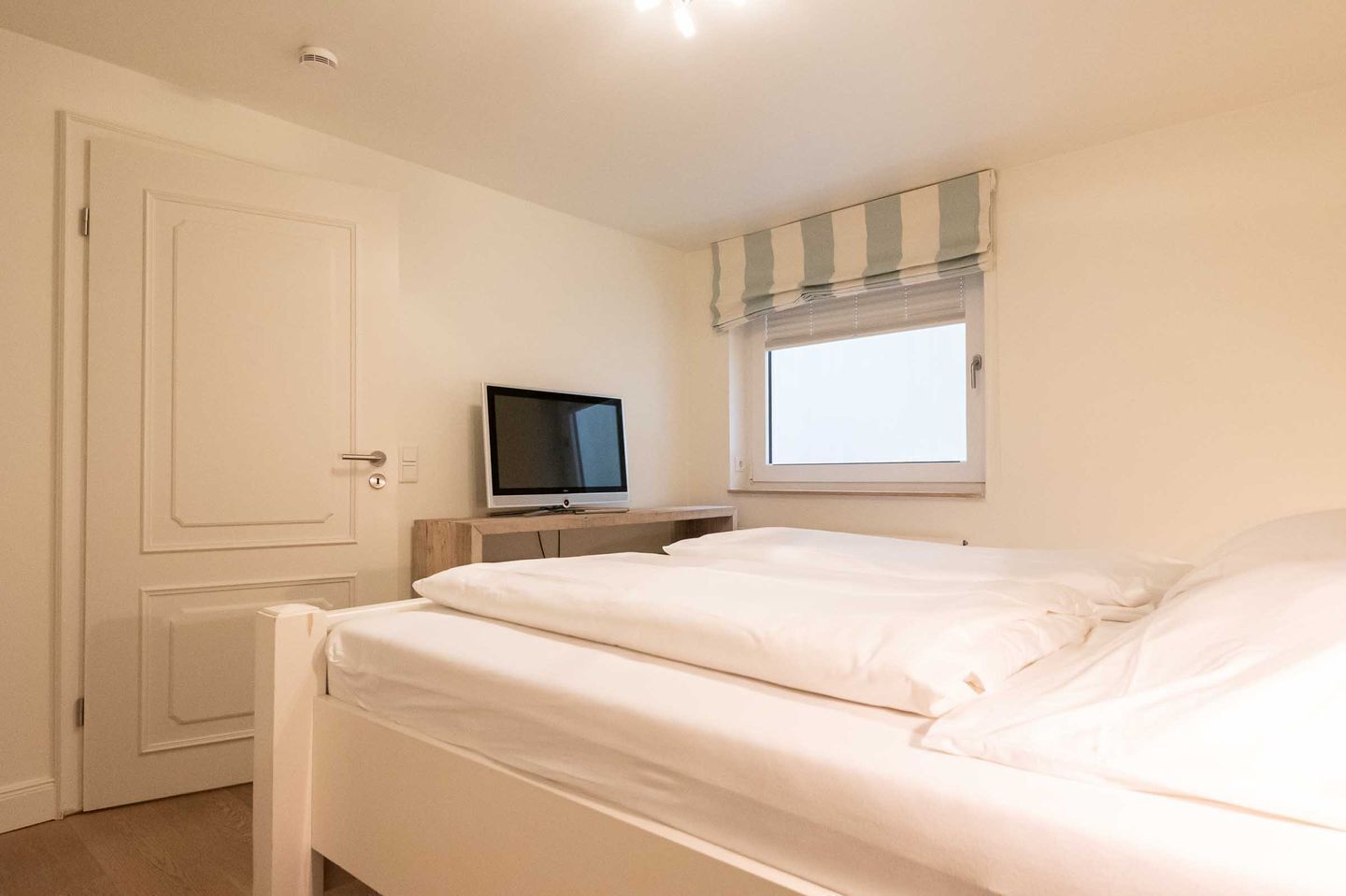 A bedroom with a bed, a television on a wooden stand, a window with blinds, and a door.