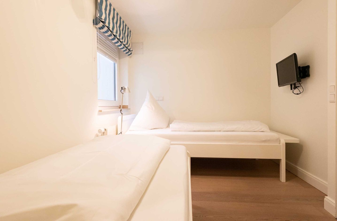 Une petite chambre à coucher au style minimaliste, avec un lit simple, une fenêtre avec des stores, un téléviseur fixé au mur et une table de chevet.
