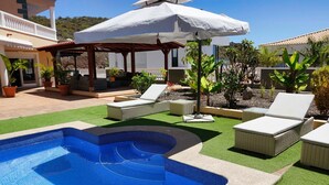 En swimmingpool med liggestole og en parasol, omgivet af krukkeplanter og en bygning med altan.