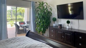 Une chambre à coucher avec un lit, un téléviseur, une grande plante en pot et une vue sur l’extérieur.