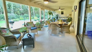 Un patio cubierto con mobiliario de mimbre, una mesa y sillas. Amplias ventanas ofrecen vistas al exterior.