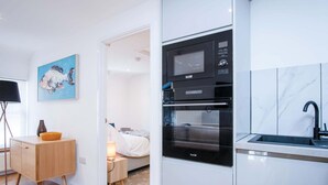 A compact kitchen with a built-in oven and microwave, a small bedside table with a lamp, and a painting on the wall.