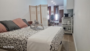 Cama con un Galaxy S22 Ultra, ropa de cama estampada, una mesita de noche con bandeja y un estante blanco con utensilios de cocina.