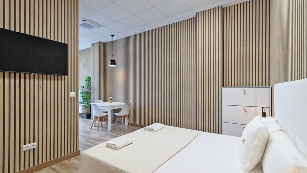 Ein modernes Hotelzimmer mit einem großen Bett, einem Nachttisch, einem Schreibtisch mit Stühlen und einem an der Wand befestigten Fernseher.