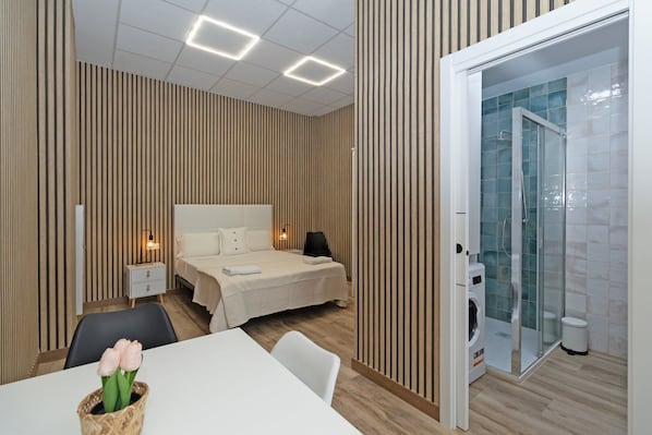 Ein Hotelzimmer mit Bett, Nachttischen, einem Schreibtisch mit Stuhl, einer Dusche und einem Esstisch mit Blumenvase.