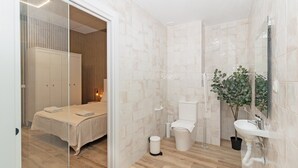 A modern bathroom with a shower, toilet, and sink. There is a bed visible through a glass door in the adjacent room. - GuestReady Essential- Blissful stay near the beach (Valencia)