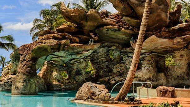A pool area with a natural rock formation, palm trees, and a clear blue sky. - Disney Aulani Two Bedroom Poolside Villa! (Kapolei)