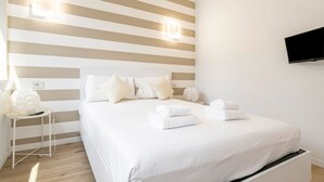 Una camera d'albergo con una parete a righe, un letto con lenzuola bianche, un comodino con una lampada e una TV a parete.